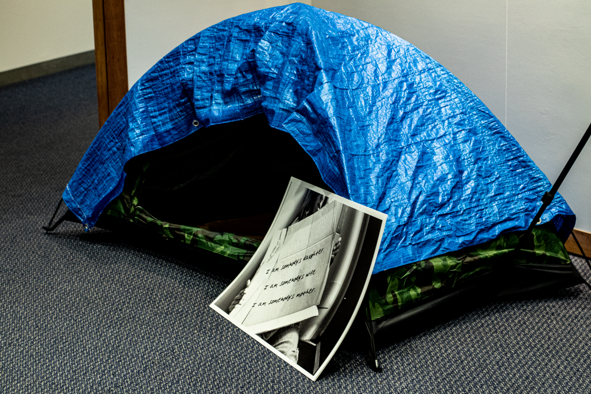 A blue tarp in the shape of a small tent lies on the ground in front of a poster offering resources for homeless people as part of the Blue Tarp Project exhibit by artist Virginia Becker. Gia Pham | Spartan Daily